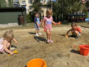 Dziewczynka stoi w środku piaskownicy trzymając grabki w ręce. W tle widać pozostałe dzieci nasypujące piasek do wiaderek.