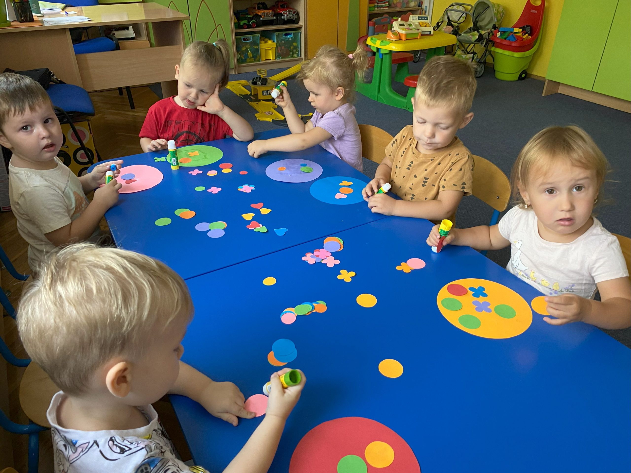 Dzieci siedzą przy stolikach na których leżą powycinane z kolorowego papieru ozdoby (kwiatuszki, kółka,serduszka)