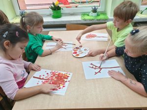 Troje dzieci siedzi przy stoliku. Malują jesienne drzewo- stemplując palcami po kartce, zamoczonymi w farbie.