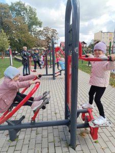 Dwie dziewczynki na siłowni plenerowej. jedna próbuje się unosić na drążkach, druga ćwiczy "wiosłowanie".