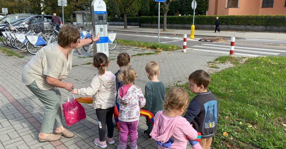 Nauczycielka przed przejściem dla pieszych tłumaczy dzieciom zasady bezpiecznego przechodzenia przez jezdnię