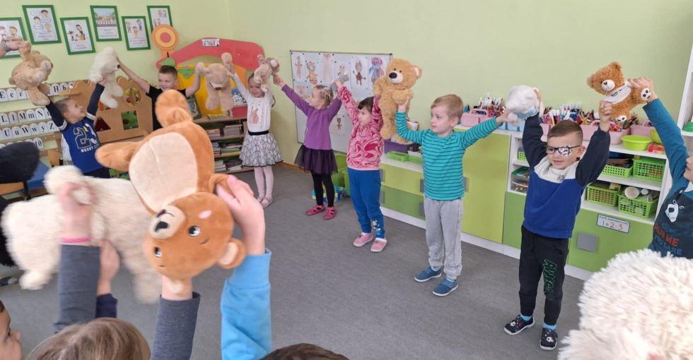 Dzieci stoją w kole. Między nimi są pluszowe misie, które trzymają. Wszyscy mają ręce w górze.