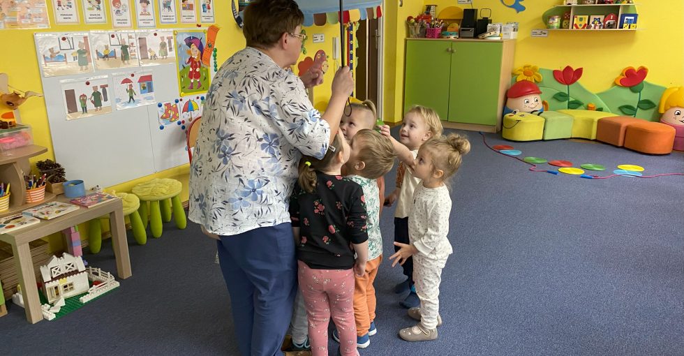 nauczycielka trzyma w ręce nad głową parasol , a pod nim gromadzą się dzieci