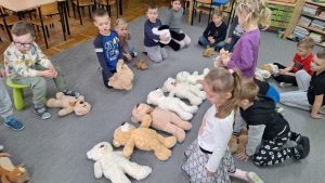 Dzieci siedzą w kole. Część z nich trzyma w rękach pluszowe misie. Pozostałe pluszaki leżą w środku sali. Dzieci wskazują największe z nich.