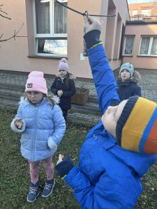 Chłopiec zawiesza smakołyki dla ptaków na gałęzi drzewka.
W tle widać 3 dzieci czekających na swoją kolej.