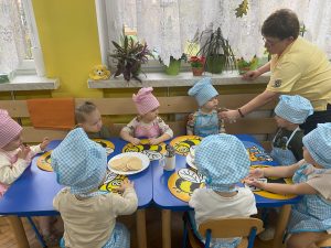 dzieci siedzą przy stolikach ubrane w czapkę i fartuch kucharski, nauczycielka daje każdemu powąchać miód ze słoika