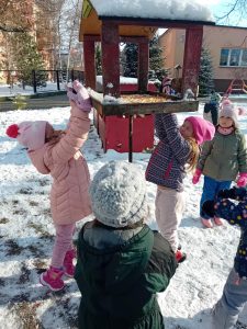 Dzieci wsypują ziarna kukurydzy do karmnika w ogrodzie przedszkolnym.