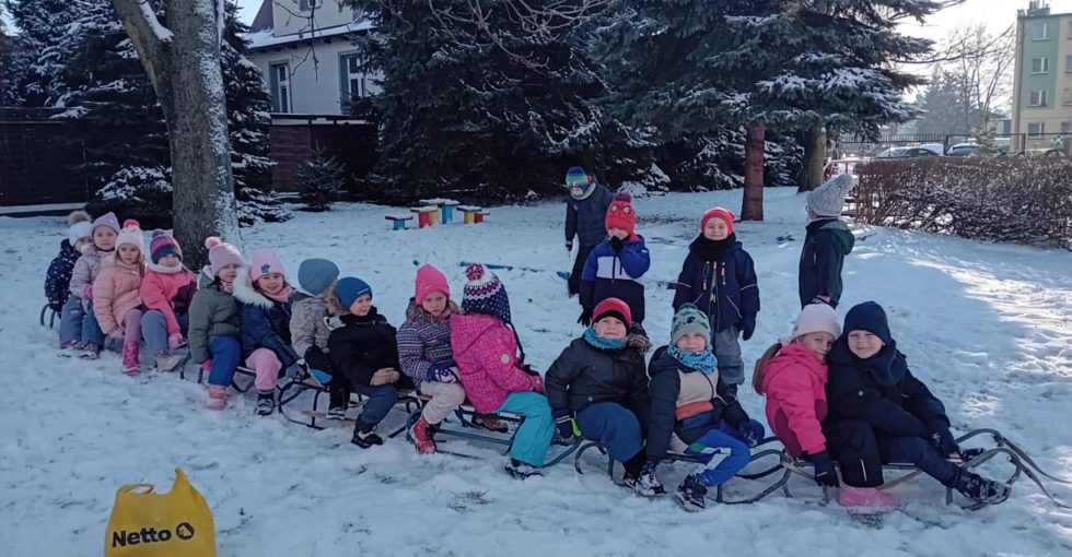 Dzieci pozują do zdjęcia, siedząc na sankach w ogrodzie przedszkolnym.