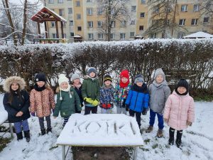 Dzieci z grupy V pozują do wspólnego zdjęcia. Przed nimi jest zasypany śniegiem stolik na którym widnieje napis KOTKI.