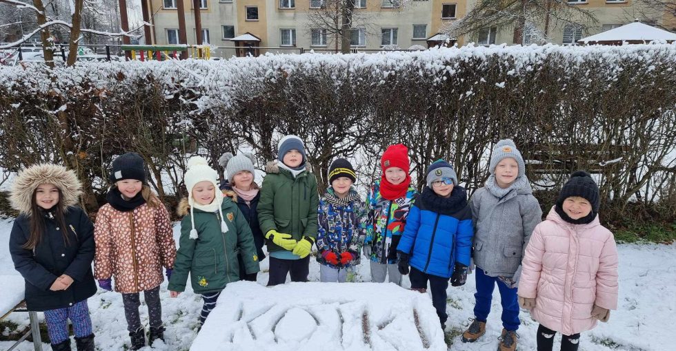 Dzieci z grupy V pozują do wspólnego zdjęcia. Przed nimi jest zasypany śniegiem stolik na którym widnieje napis KOTKI.