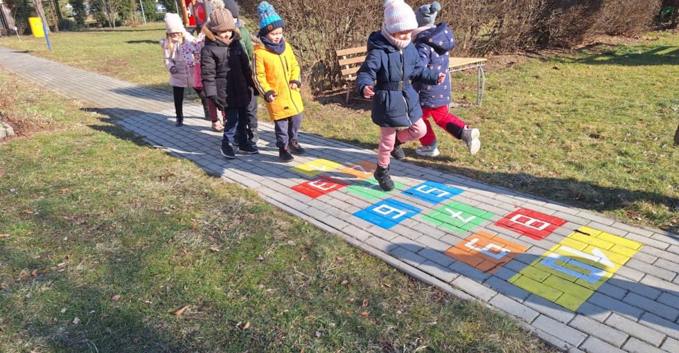 Grupa dzieci stoi w kolejce, jedno z nich skacze po namalowanych na chodniku klasach.